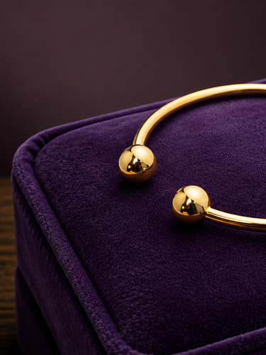 A polished gold cuff bracelet with two small spherical bead details, displayed on a purple velvet jewelry box against a dark background.