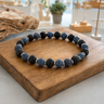 A beaded stretch bracelet made of alternating black lava stone and matte blue agate marbled beads, separated by small steel spacer beads, displayed on a wooden block.