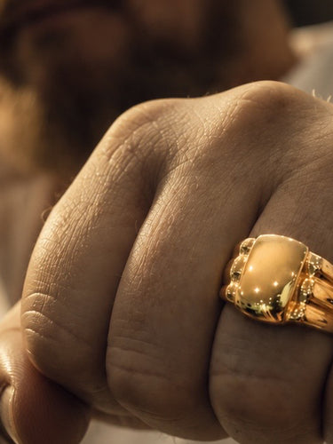 A close-up of a man's hand wearing a large bespoke gold signet ring with a polished oval center stone, set against a dark, blurred background.